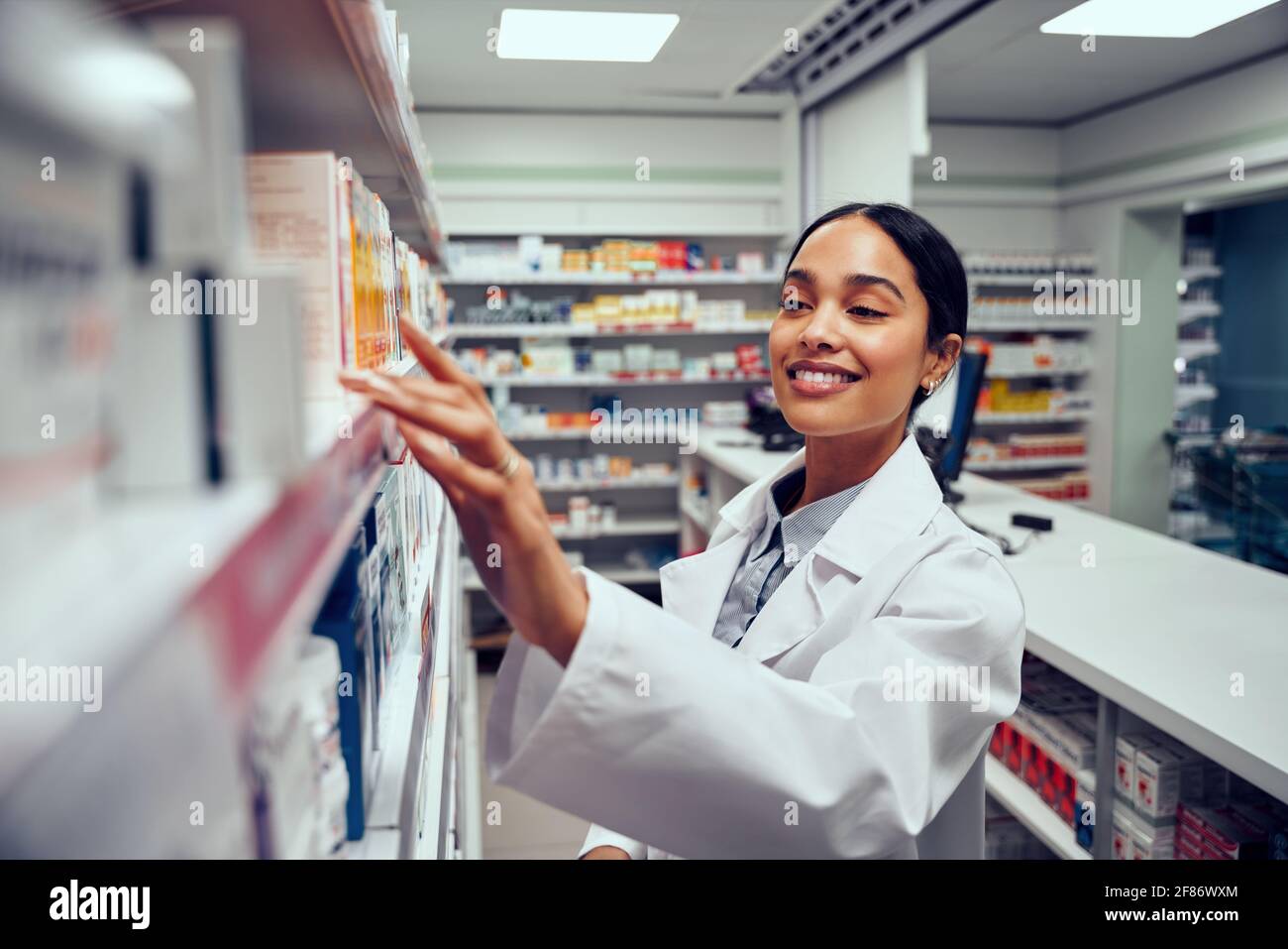 Sorridente giovane farmacista femminile che indossa un camice da laboratorio in piedi dietro il bancone guardando per la medicina in scaffale Foto Stock