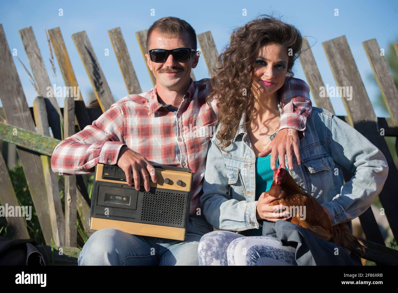 Una ragazza con un pollo e un uomo con un magnetofono retrò sono seduti sulla panchina. Gli abitanti del villaggio sono nello stile degli anni novanta. Foto Stock