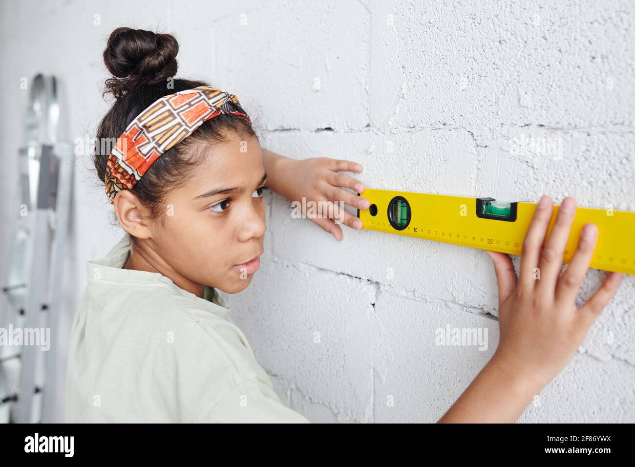 Serious African femmina adolescente in piedi da muro di mattoni dipinto bianco e usando l'attrezzo manuale di misura di livello durante il lavoro domestico di rinnovamento Foto Stock
