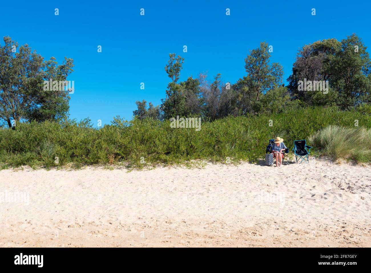 Una donna si siede da sola leggendo un libro sotto un grande Cappello da spiaggia su una sedia da spiaggia sulla spiaggia principale di Merimbula Sulla costa sud del nuovo Galles del Sud dell'Australia Foto Stock