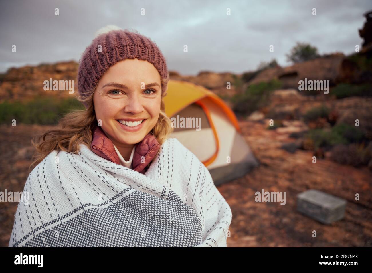 Donna sorridente con coperta sulla montagna in piedi fuori tenda durante campeggio in collina di montagna Foto Stock