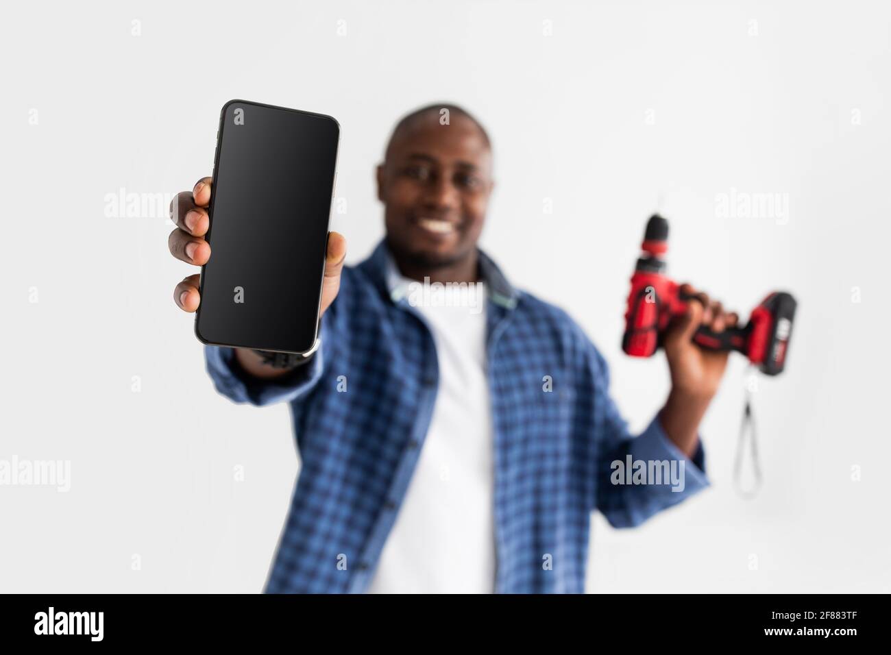 Felice handyman afroamericano che tiene il cacciavite elettrico e mostra il telefono con schermo vuoto su sfondo bianco Foto Stock
