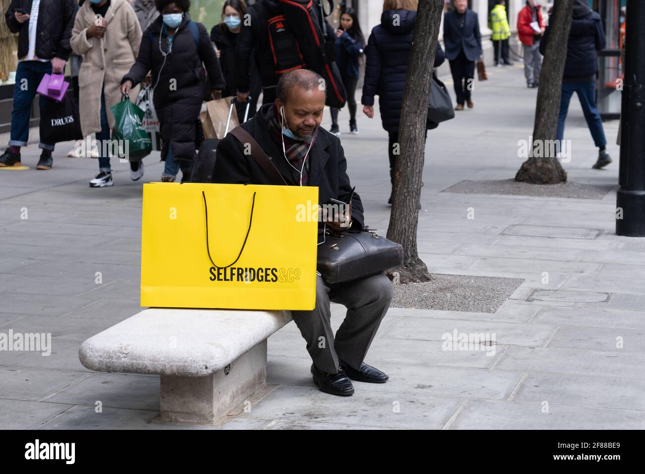 Londra, Inghilterra, 12 aprile 2021. Uomo con grande borsa Selfridges giallo seduto in Oxford Street come Covid restrizioni sono attenuate permettendo negozi non essenziali di aprire. Fotografo : Brian Duffy Foto Stock