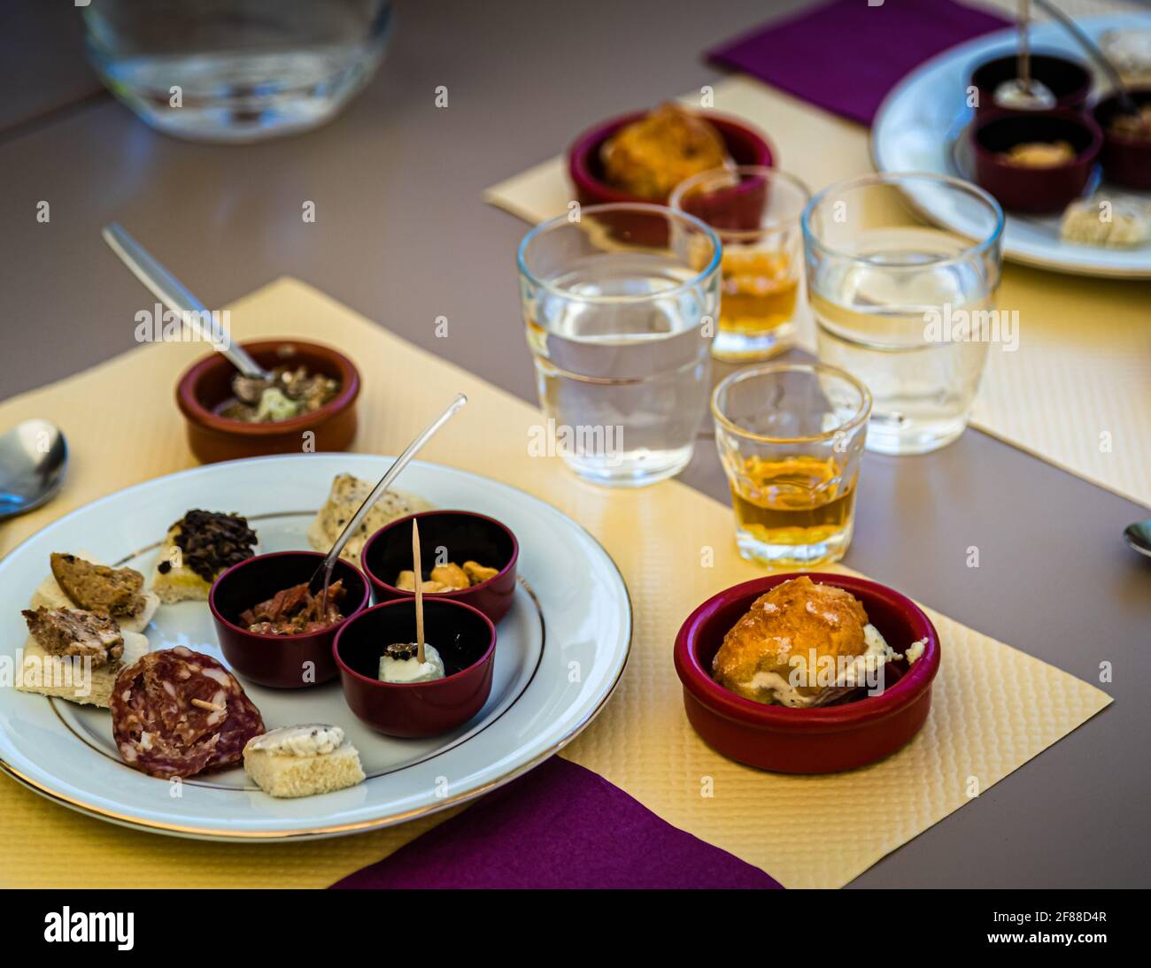 Un piatto di degustazione vario con salame al tartufo e altri piccoli piatti, forse compresi rillettes al tartufo, è posato sul tavolo alla La Maison aux Mille Truffes a Marey-lès-Fussey in Borgogna, Francia, accanto a bicchieri d'acqua e due piccoli bicchieri di uno spirito giallo-dorato. I visitatori della tenuta possono aggiungere una tale degustazione al loro tour – la gamma va dal salame al tartufo e dal fagiano con tartufo a un gelato al tartufo servito in pasta choux. Foto Stock