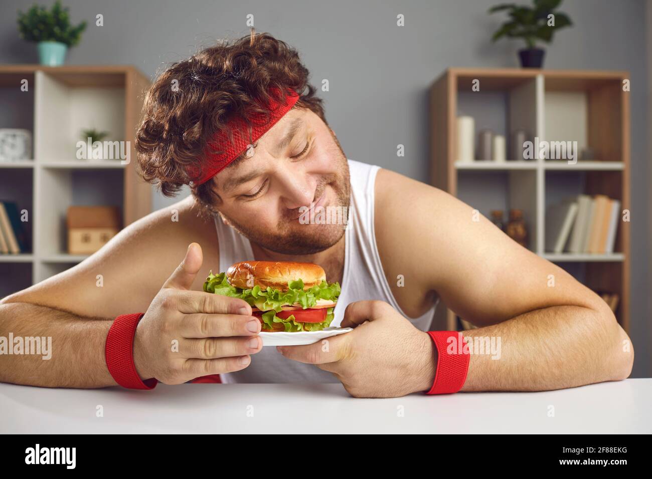 Chubby obese uomo caucasico in abbigliamento sportivo che tiene hamburger su piatto verticale Foto Stock