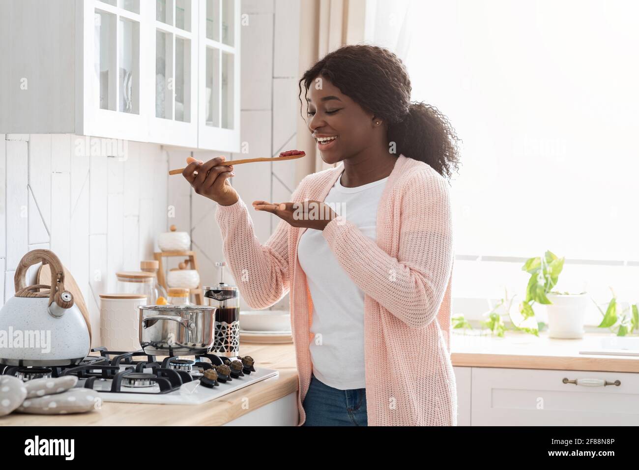 Cheerful Black Housewife degustazione cibo mentre cucinare sano pranzo in Cucina moderna Foto Stock