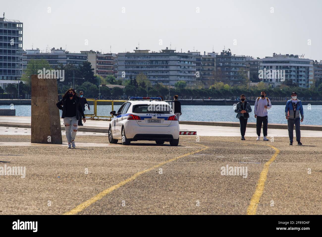 Salonicco, Grecia - Aprile 11 2021: Polizia greca pattugliando auto accanto alla folla indossando covid-19 maschere all'aperto. Persone con protezione del viso a piedi sulla zona pedonale del lungomare della città. Foto Stock
