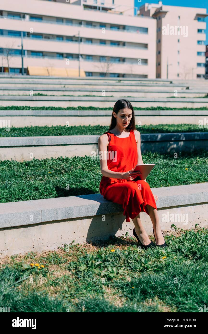 Donna caucasica che lavora con il suo tablet. È seduta nel campus nelle scale con erba verde. Abito rosso Foto Stock