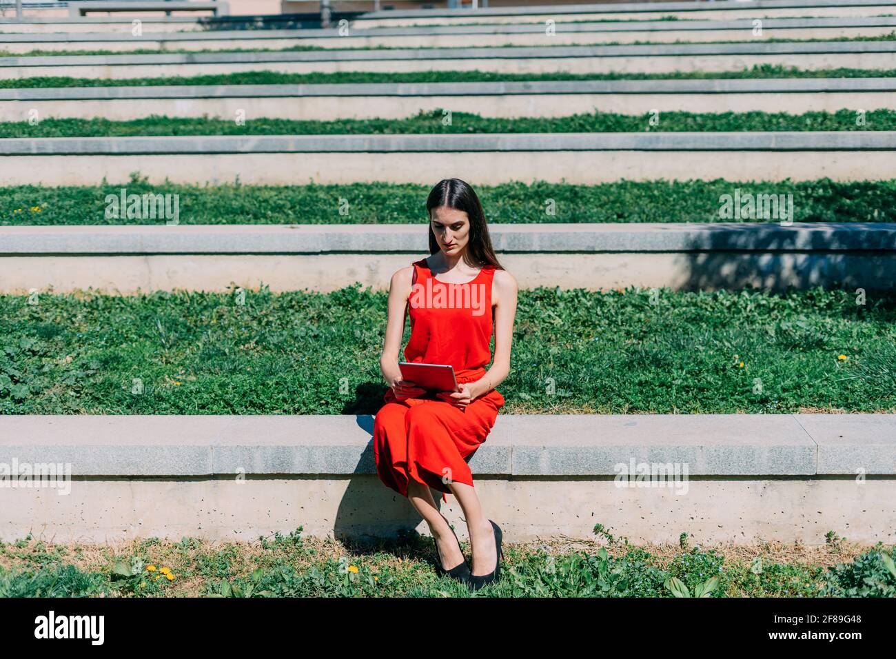 Donna caucasica che lavora con il suo tablet. È seduta nel campus nelle scale con erba verde. Abito rosso Foto Stock