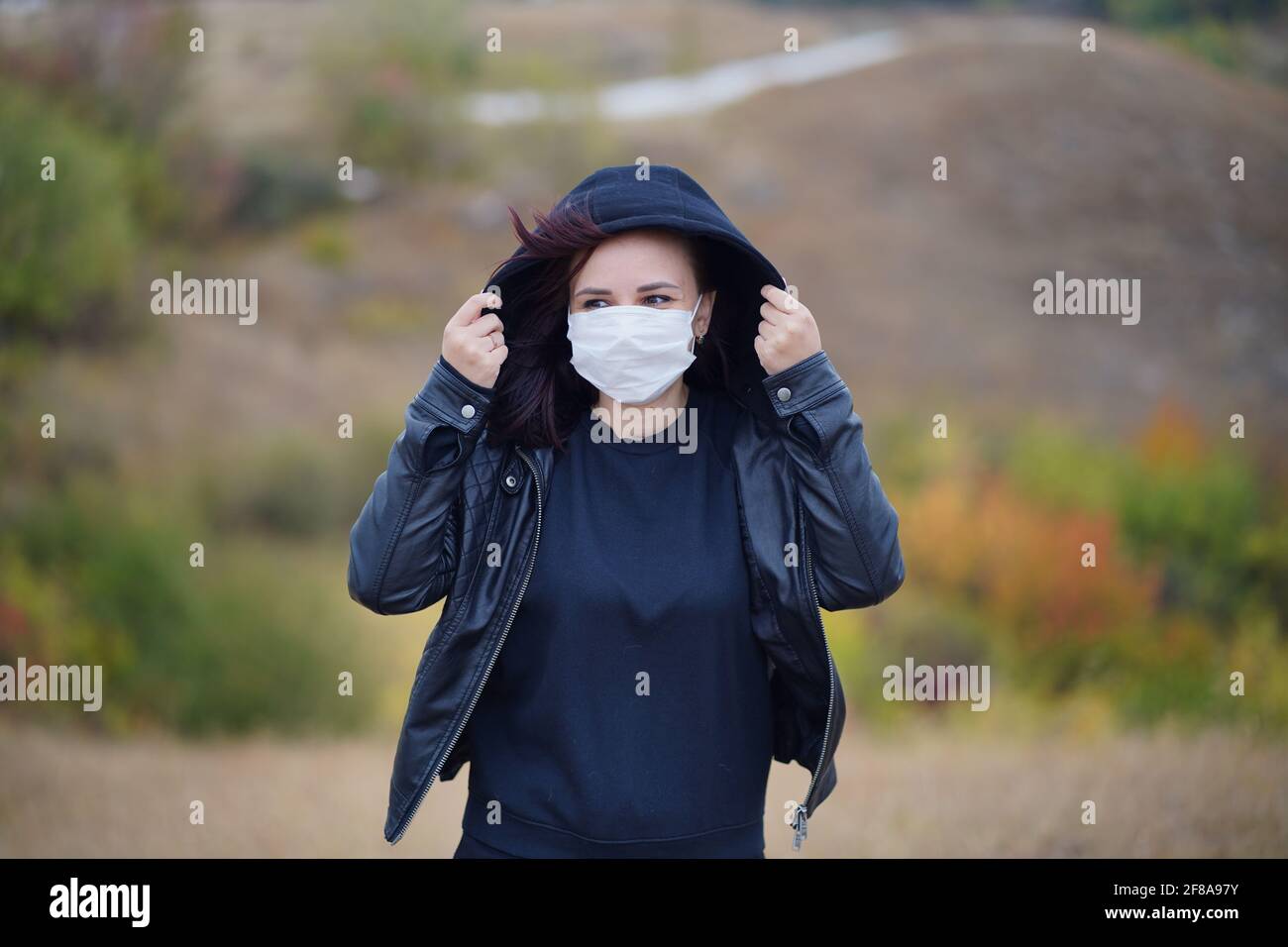 Giovane donna in maschera medica in piedi in campagna. Brunette per adulti in maschera protettiva in tempo fresco a piedi su terreno collinare Foto Stock