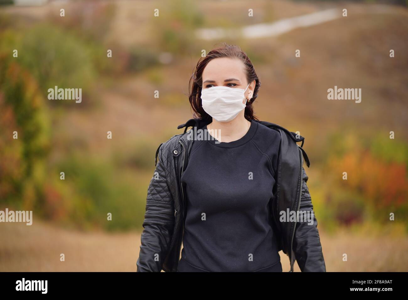 Giovane donna in maschera medica in piedi in campagna. Brunette per adulti in maschera protettiva in tempo fresco a piedi su terreno collinare Foto Stock