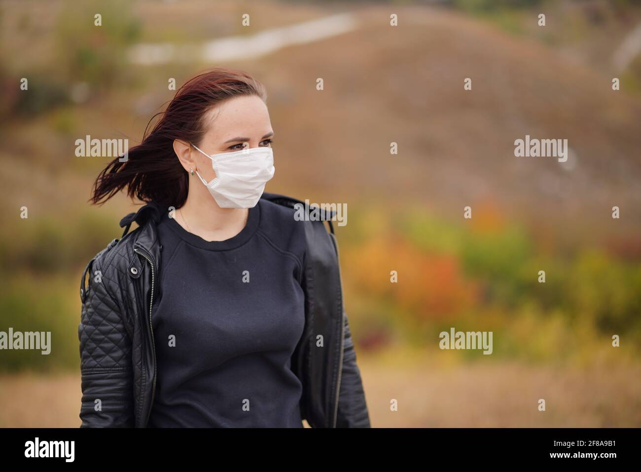 Giovane donna in maschera medica in piedi in campagna. Brunette per adulti in maschera protettiva in tempo fresco a piedi su terreno collinare Foto Stock
