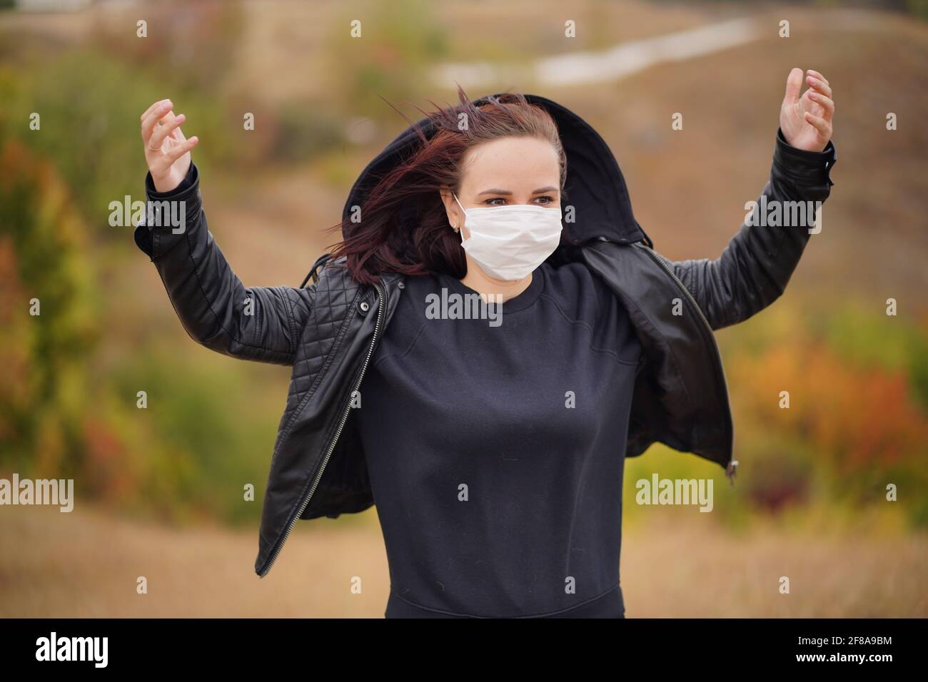 Giovane donna in maschera medica in piedi in campagna. Brunette per adulti in maschera protettiva in tempo fresco a piedi su terreno collinare Foto Stock