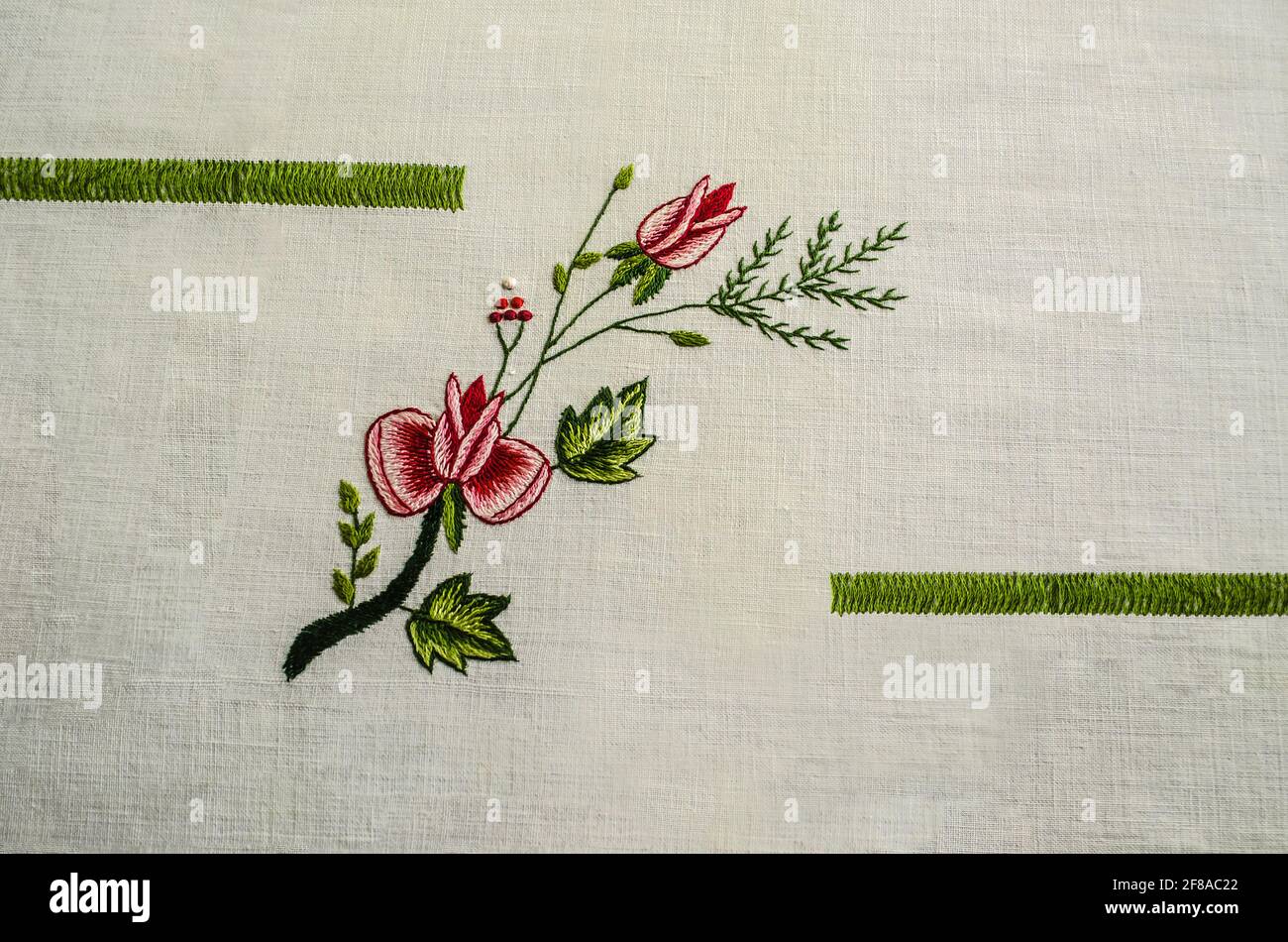 Ricamato tra nastri verdi, un rametto con rose rosse e un bocciolo, intrecciato con un rametto di conifere e bacche rosse su un tessuto di cotone bianco Foto Stock