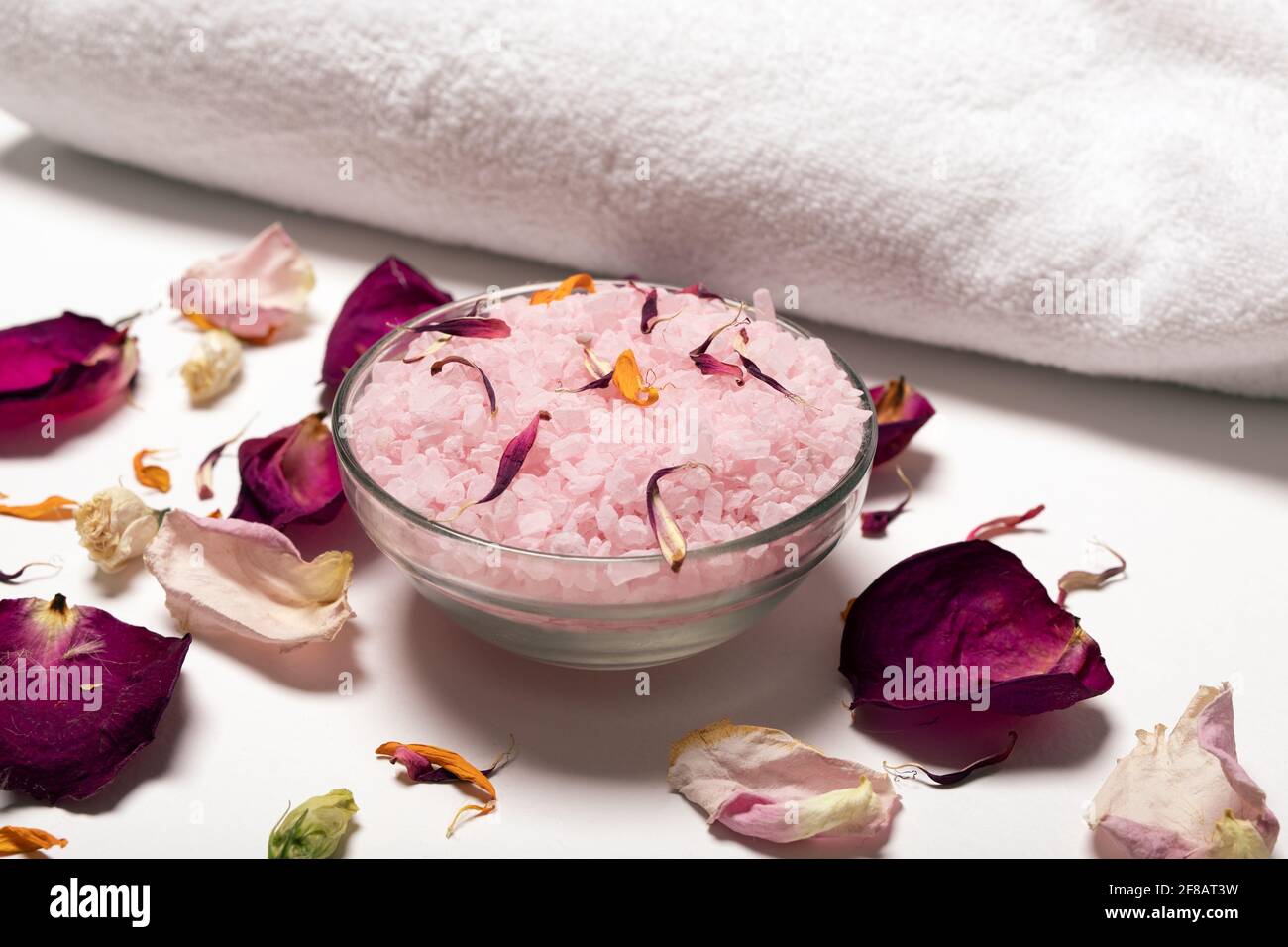 Immagine di tenerezza di vaso di vetro riempito di sale rosa circondato da petali di rosa e asciugamano bianco su superficie bianca. Trattamenti Spa e relax o di massa Foto Stock