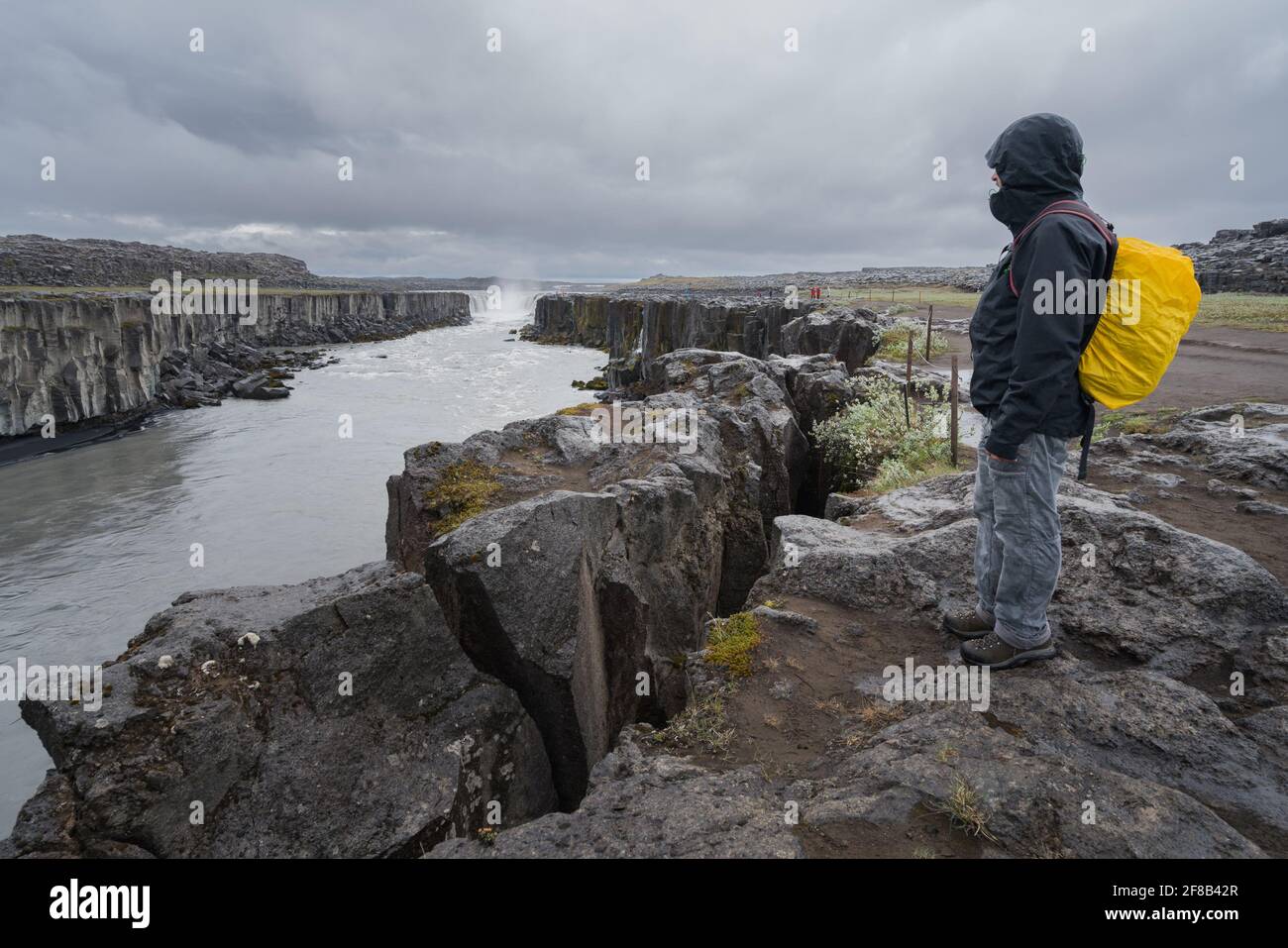 Uomo in abiti all'aperto con zaino giallo in piedi su una scogliera rocciosa, con vista sulla cascata Selfoss in Islanda in una giornata nuvolosa. Foto Stock