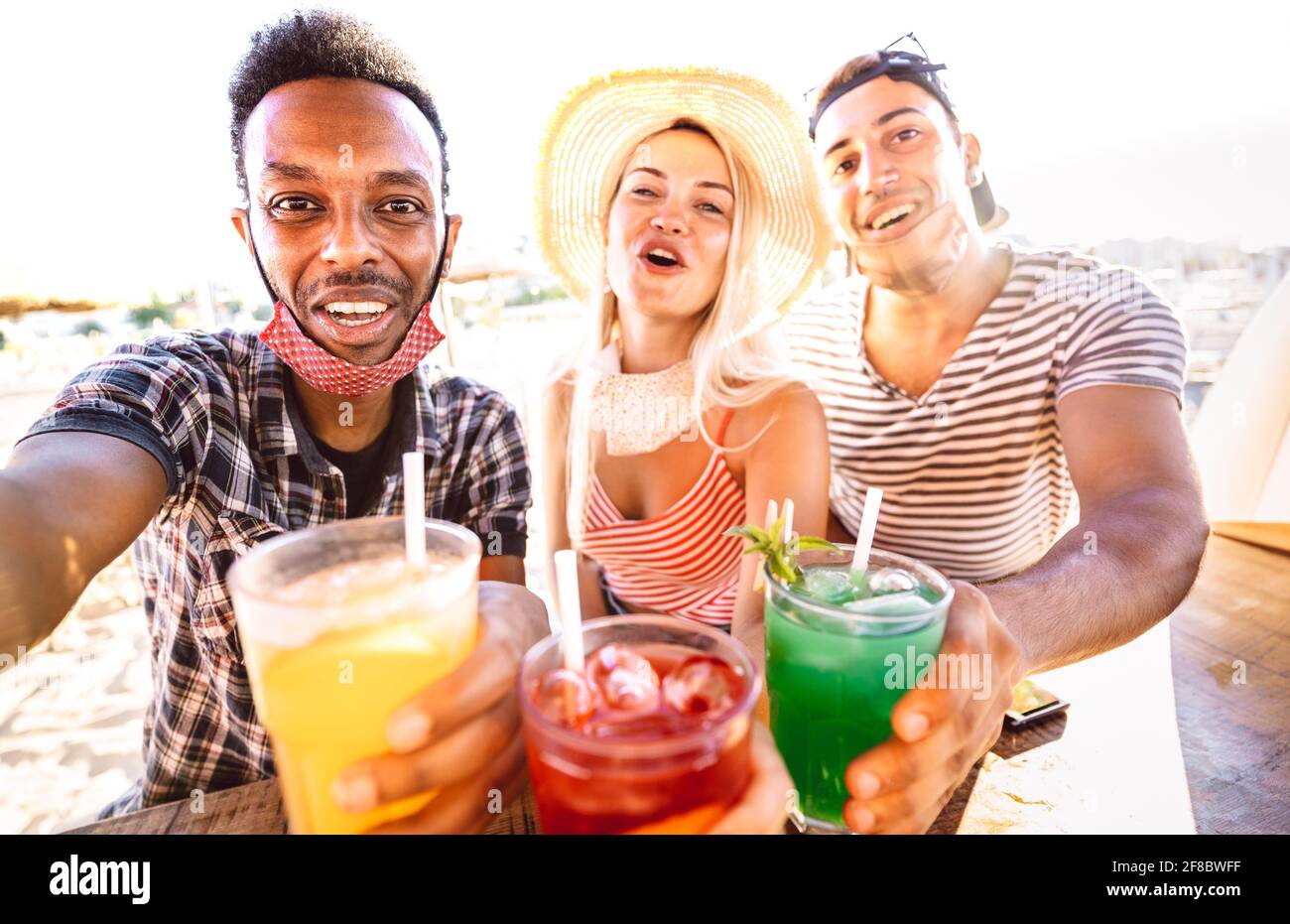 Trio di persone multirazziali che prendono selfie con maschere aperte al viso beach bar - nuovo concetto di vita normale con ubriaco amici che si divertono insieme Foto Stock