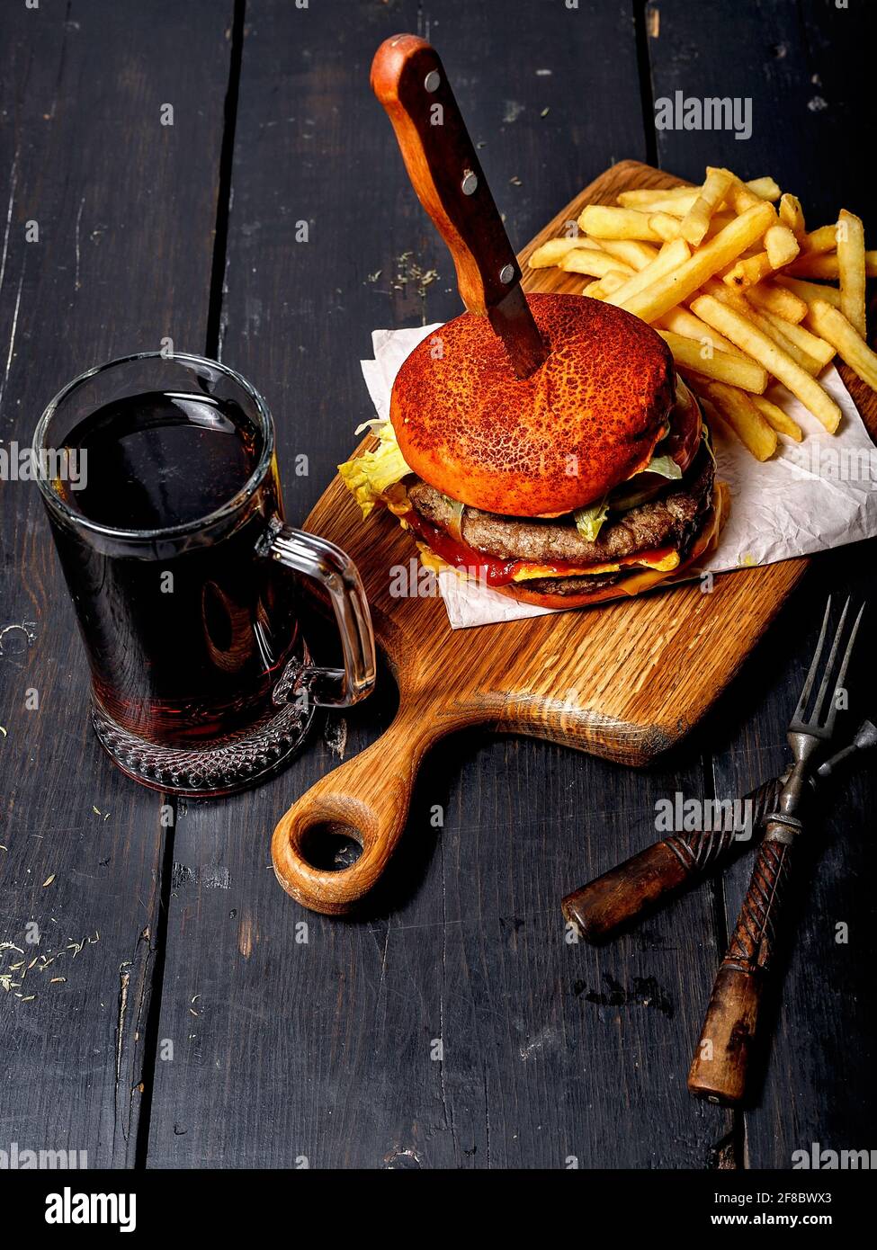 Appetitoso hamburger di manzo doppio e patate fritte su un tagliere di legno, e una tazza di birra scura. Rustico sfondo scuro. Fotografia di fast food. V. Superiore Foto Stock