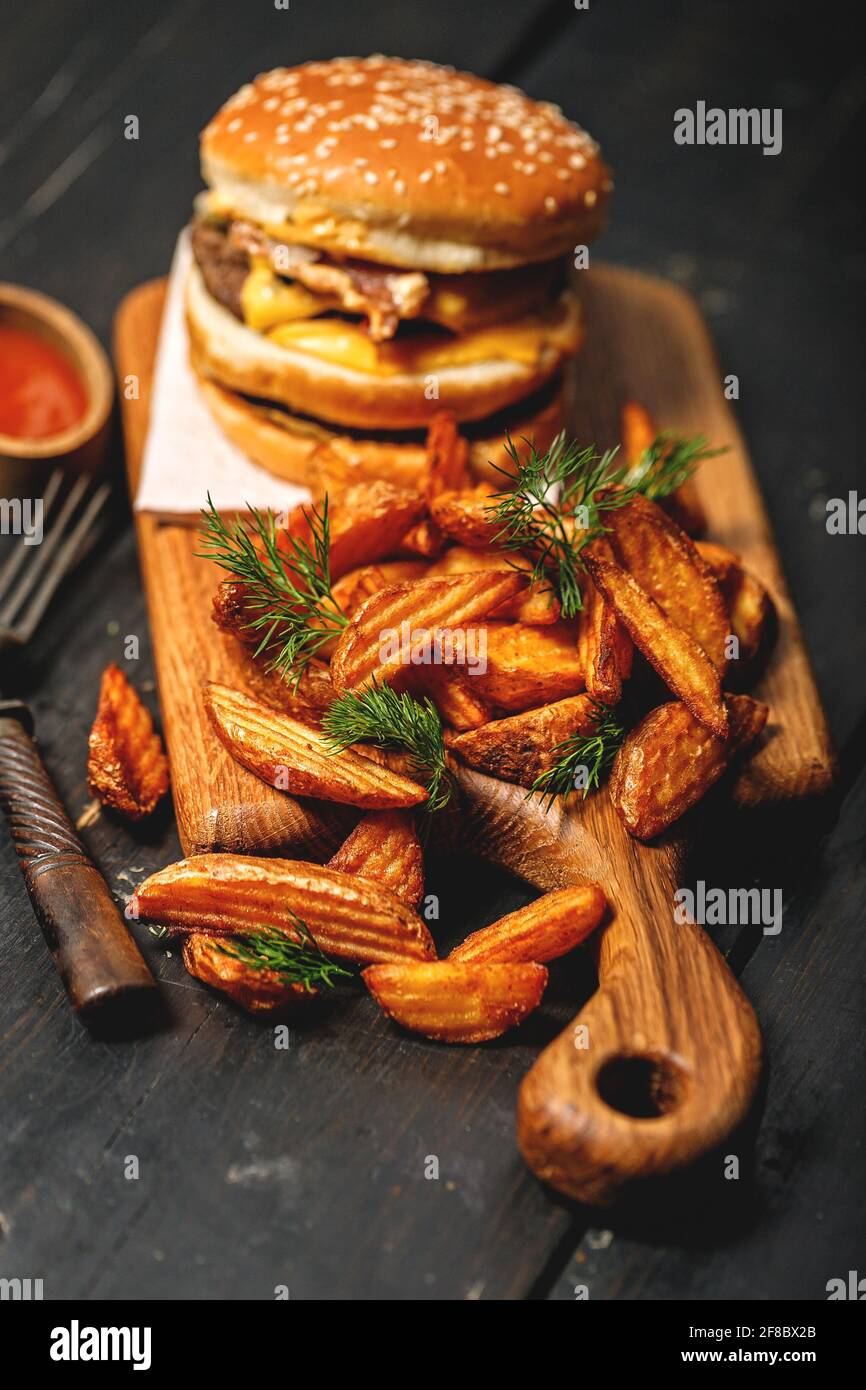 Appetitoso hamburger di manzo doppio e una porzione di patatine fritte su un tagliere di legno. Rustico sfondo scuro. Fast food fotografia di natura morta. Sho. Verticale Foto Stock