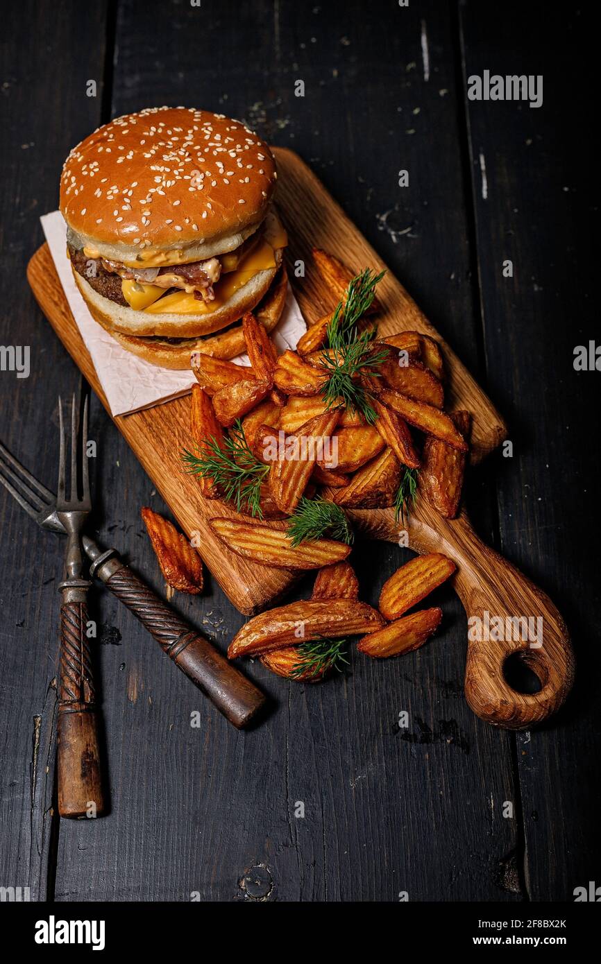 Fette di patate fritte con erbe e un delizioso doppio hamburger su un tagliere di legno. Rustico sfondo scuro. Fotografia di fast food. Shot verticale Foto Stock