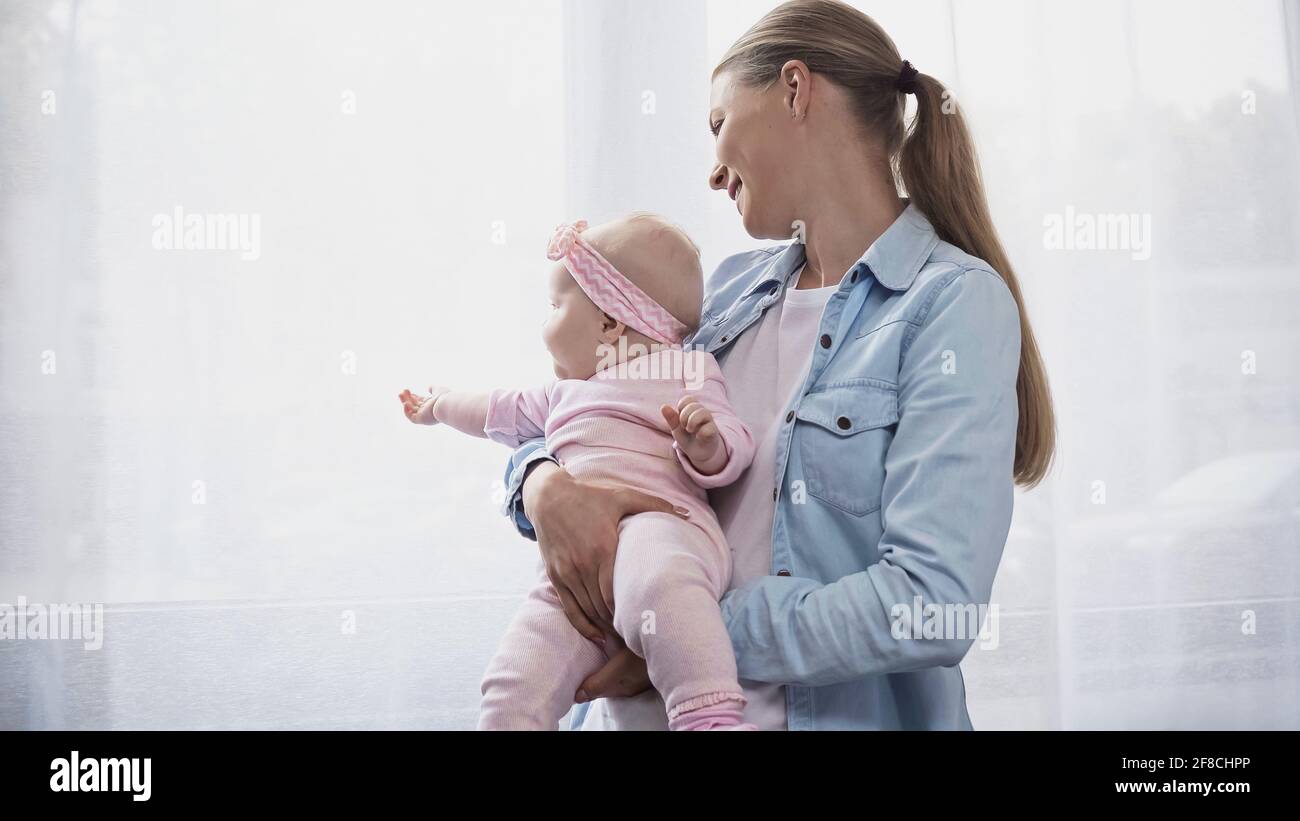 madre allegra che tiene in armi figlia del bambino che punta con la mano alla finestra Foto Stock