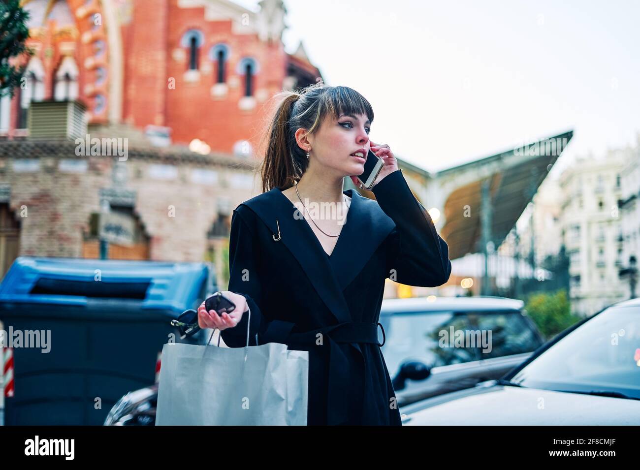 Ragazza giovane alla moda in abito nero con borse per la spesa è parlare al telefono sulla strada della città Foto Stock