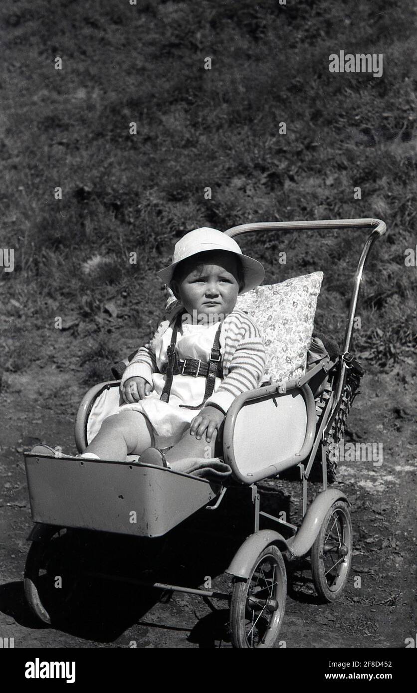 Anni '50, storico, fuori in un giorno d'estate, un bambino neonato che indossa un cappello, seduto in una passeggino con struttura in metallo o buggy dell'epoca, con poggiapiedi attaccato e un cuscino per sostegno alla schiena, Inghilterra, Regno Unito. Foto Stock