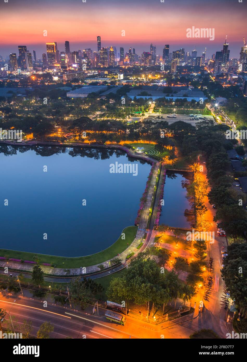 Parco pubblico Benjakiti con un lago, grattacieli e traffico a Bangkok, Thailandia al crepuscolo Foto Stock