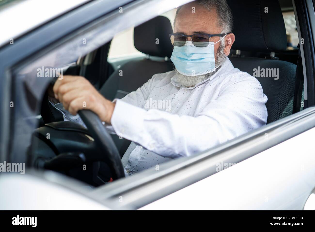 Un uomo anziano in una maschera medica che guida un'auto. Concetto pandemico di coronavirus. Viaggio su strada, viaggio e concetto di persone anziane Foto Stock