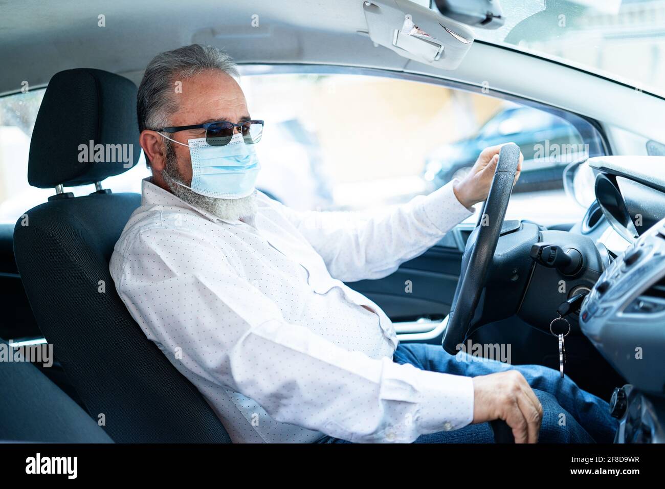 Un uomo anziano in una maschera medica chirurgica che guida un'automobile. Concetto pandemico di coronavirus. Viaggio su strada, viaggio e concetto di persone anziane Foto Stock