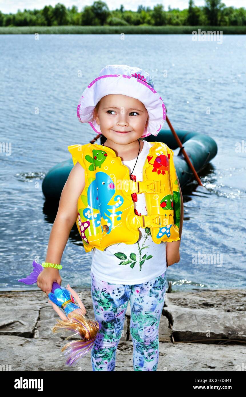 Bambina in giubbotto salvagente, in piedi sulla riva del fiume sullo sfondo di una barca di gomma. Messa a fuoco selettiva Foto Stock