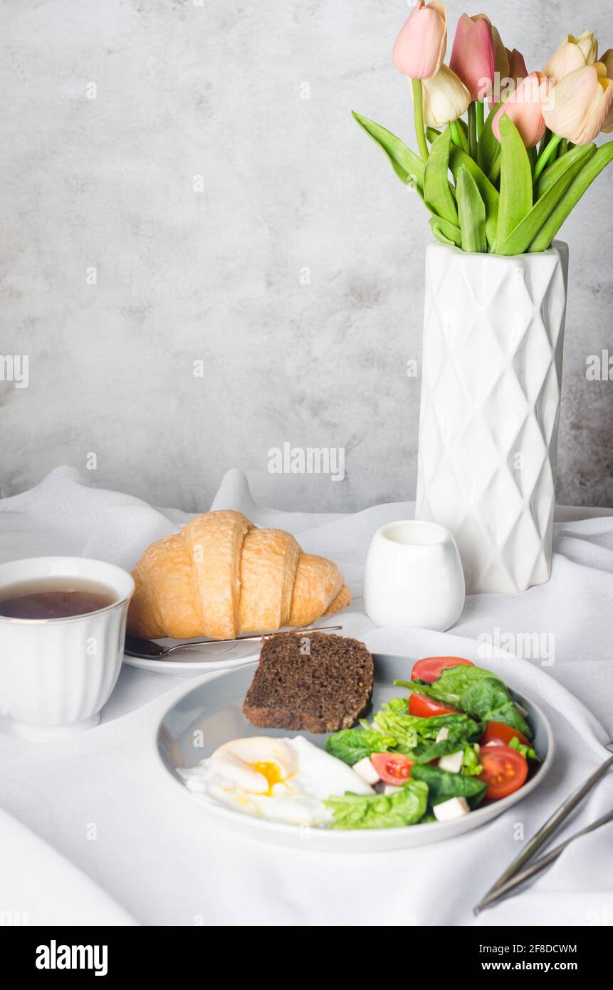 Decorazione di impostazione della tavola primaverile del mattino. Insalata in piatto, uovo, tazza di caffè e croissant, tulipani freschi in vaso su fondo tovaglia bianco pulito. Foto Stock