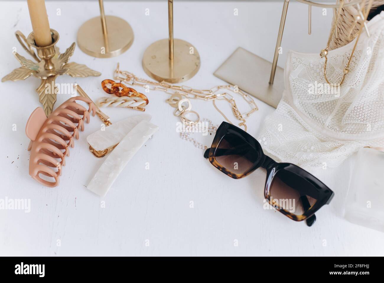 Gioielli dorati, occhiali da sole, clip per capelli, cosmetici, profumo e lingerie pizzo su tavolo bianco con candele d'epoca e specchio boho. Moderno accce estivo Foto Stock