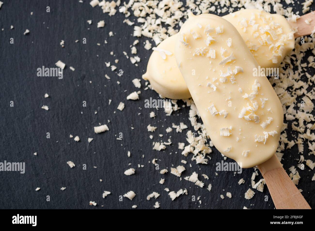 Dessert estivi e spuntini freddi con due gelaterie su un bastone o lecci, circondati da scaglie di cioccolato bianco su una pietra nera scura b Foto Stock