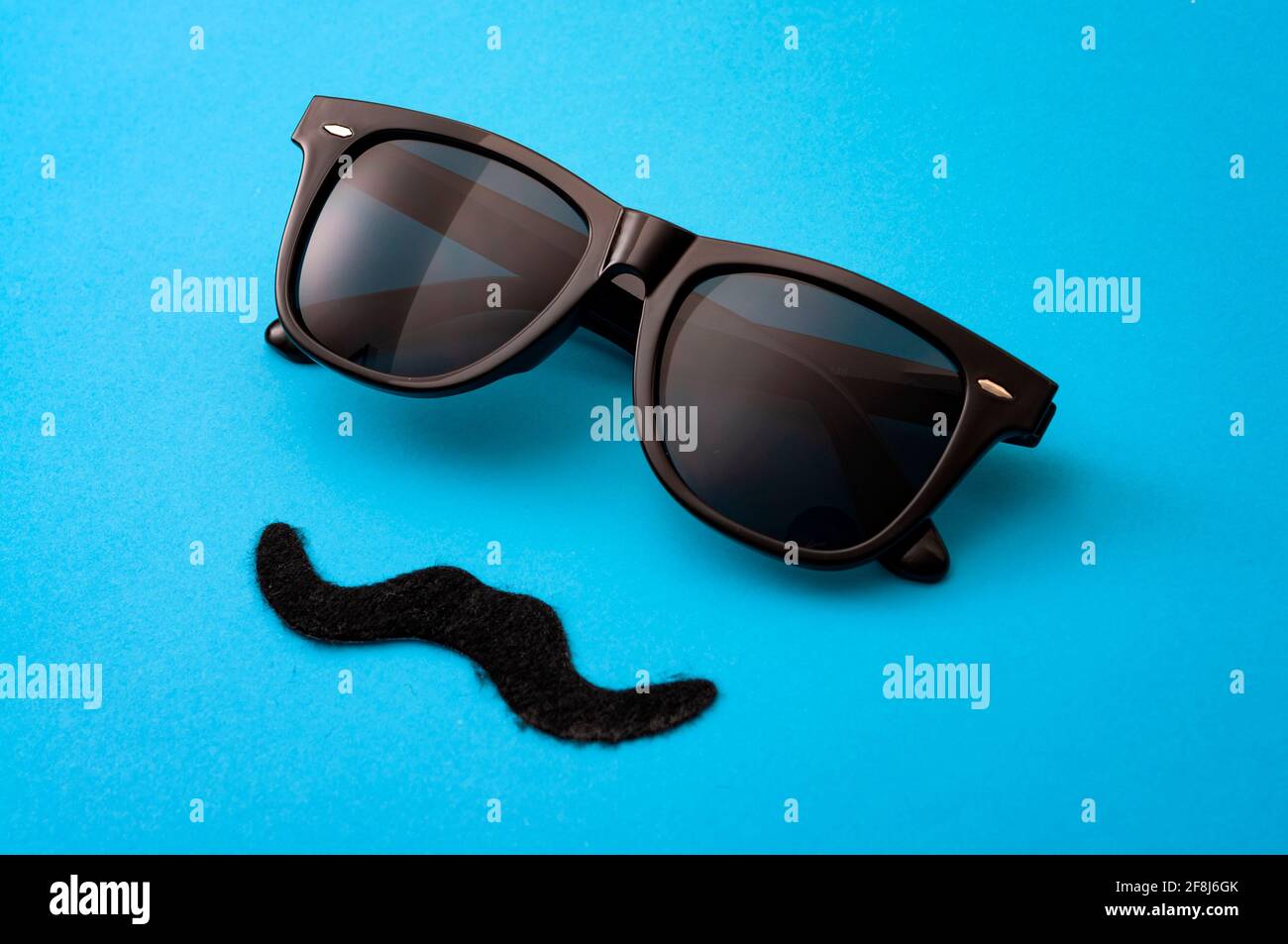 Il concetto di moda del giorno del padre e dell'hipster maschile con immagine minimalista di un paio di occhiali da sole quadrati e di un falso baffi su sfondo blu colorato Foto Stock