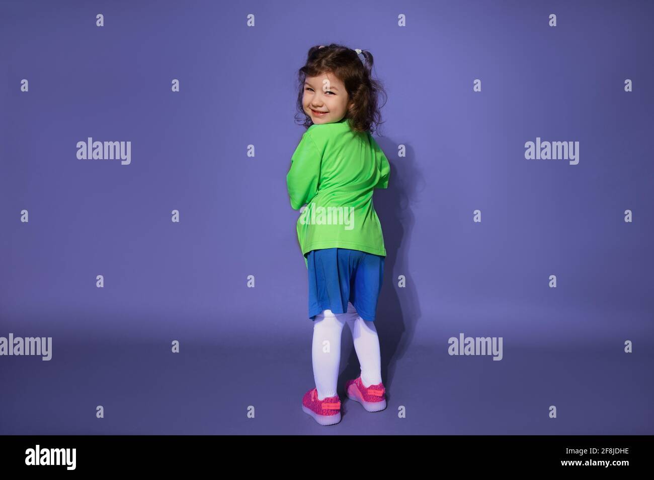 Divertente bambina in uniforme da calcio si alza con la schiena su uno sfondo viola e guarda nella cornice Foto Stock