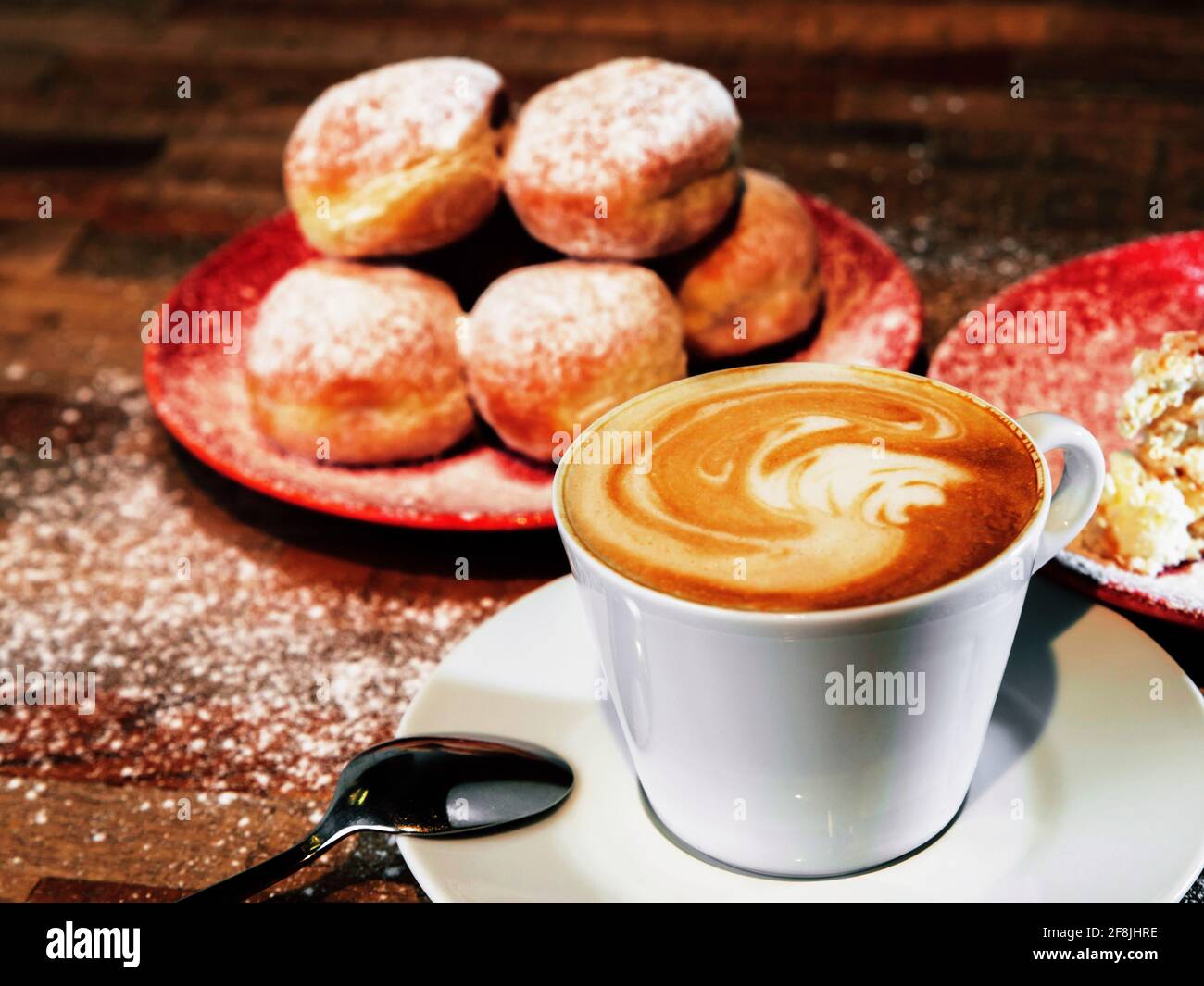 Tazza di caffè bianco piatto (espresso con schiuma di latte), gruppo di torte fritte arrotondate (ciambelle) di impasto farcite con marmellata su piatti rossi su tavolo di legno. Foto Stock