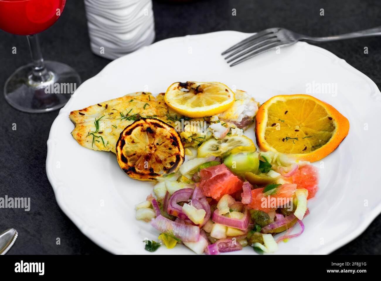 Grigliare pesce, insalata di arancia e limone con insalata di cicoria e finocchio con pomelo su piatto bianco sul tavolo con bevanda rossa e ciotola in ceramica, barbecue. Foto Stock