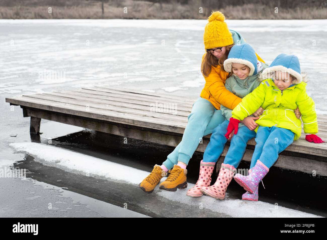 Mamma con i bambini siede su un ponte di legno da un lago ghiacciato Foto Stock