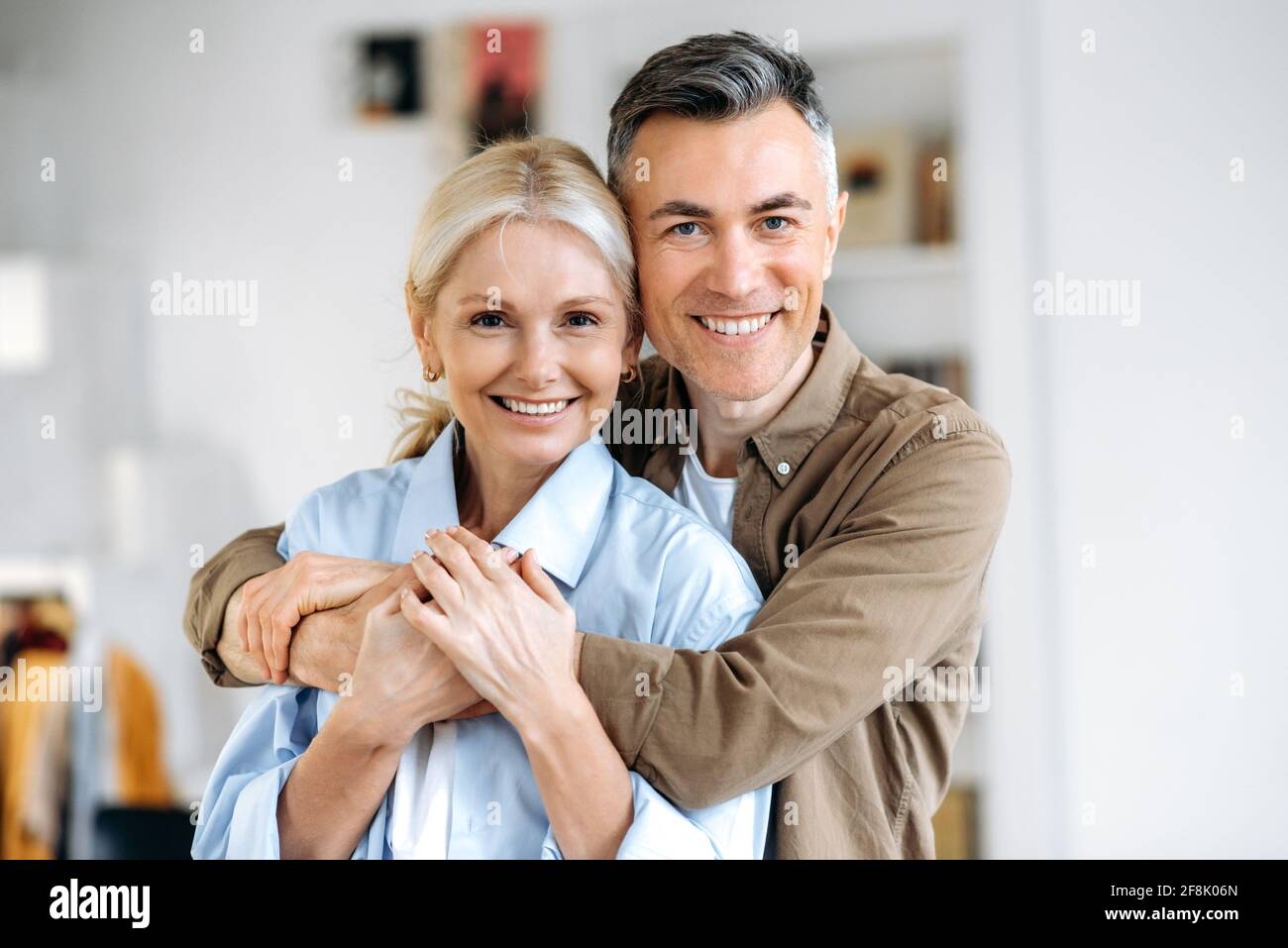 Primo piano ritratto di una bella coppia di matrimonio caucasica, marito felice e moglie vestiti in abiti casual elegante, in piedi a casa, abbracciando, guardando sorridente direttamente nella fotocamera Foto Stock