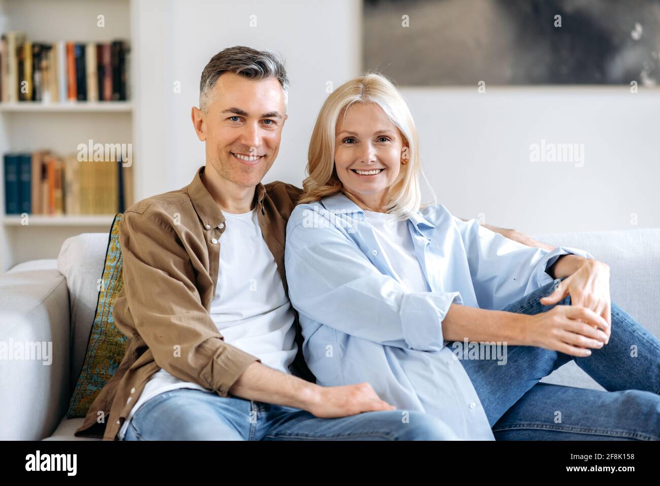 Ritratto di coppia matura innamorata. Coppie caucasiche gioiose di mezza età trascorrono il tempo insieme a casa, rilassandosi su un divano nel soggiorno, vestita con abiti casual eleganti, sorridendo alla macchina fotografica Foto Stock