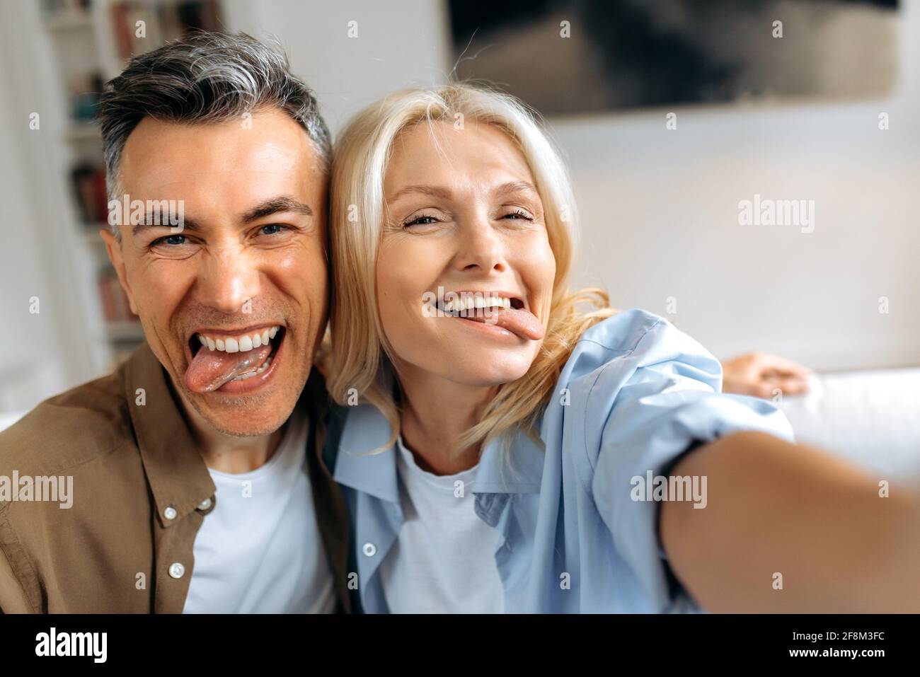 Coppia matura sposata divertente, marito caucasico di mezza età e moglie che si aggirano intorno, prendendo un selfie sugli smartphone, mostrando le loro lingue, facendo i volti, divertendosi insieme Foto Stock