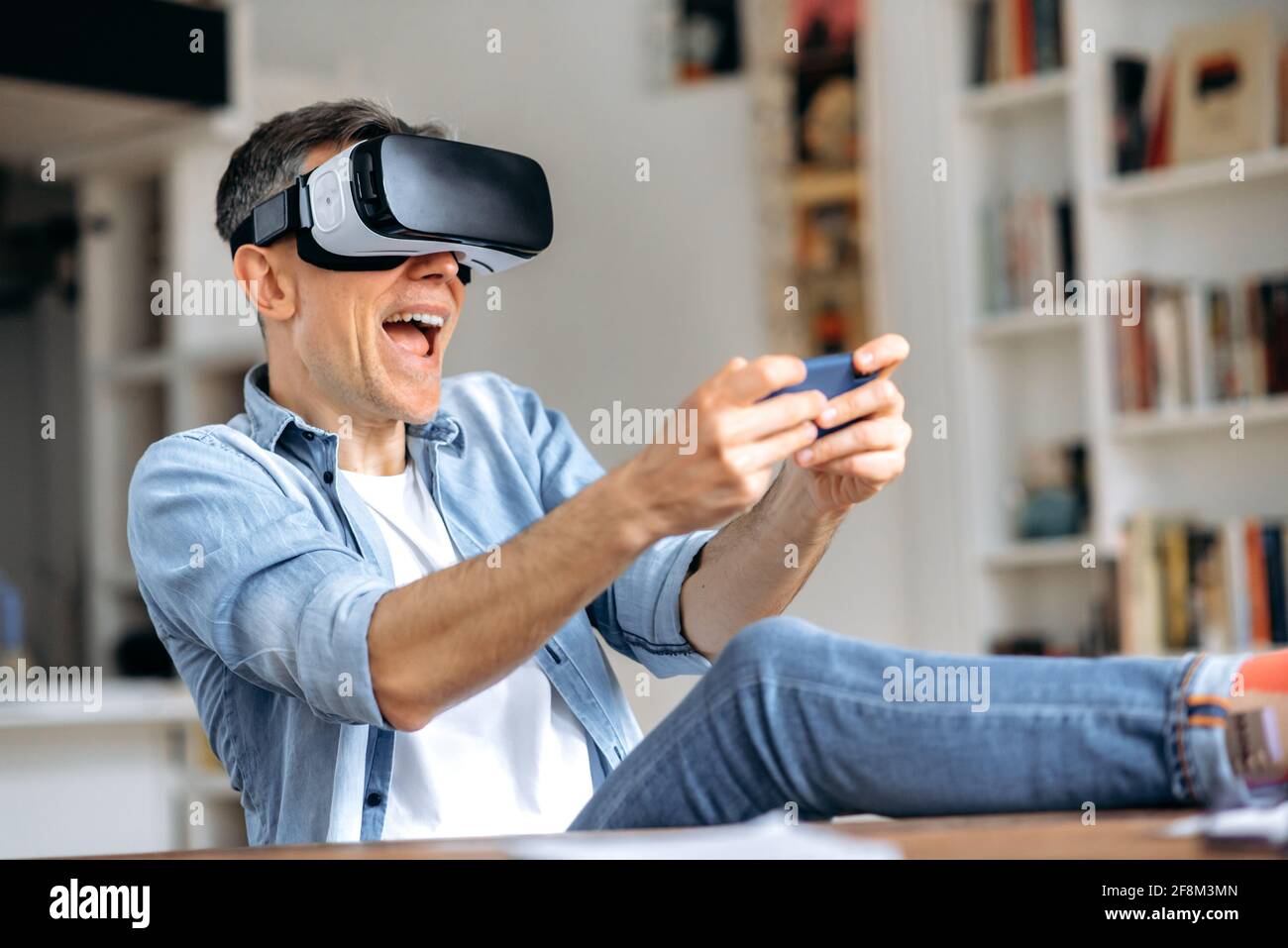 Uomo caucasico di media età, freelancer, indossando occhiali di realtà virtuale durante la pausa di lavoro. Un uomo entusiasta di testare gli occhiali VR mentre si siede alla scrivania, lo smartphone con il visore VR, la realtà virtuale Foto Stock