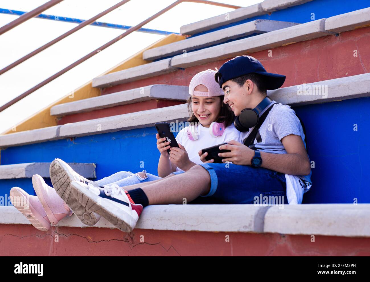 Due pre-adolescenti caucasici, molto sorridenti, con dispositivi mobili e cuffie, mostrando i loro telefoni cellulari l'uno all'altro seduti sui bleachers. Foto Stock