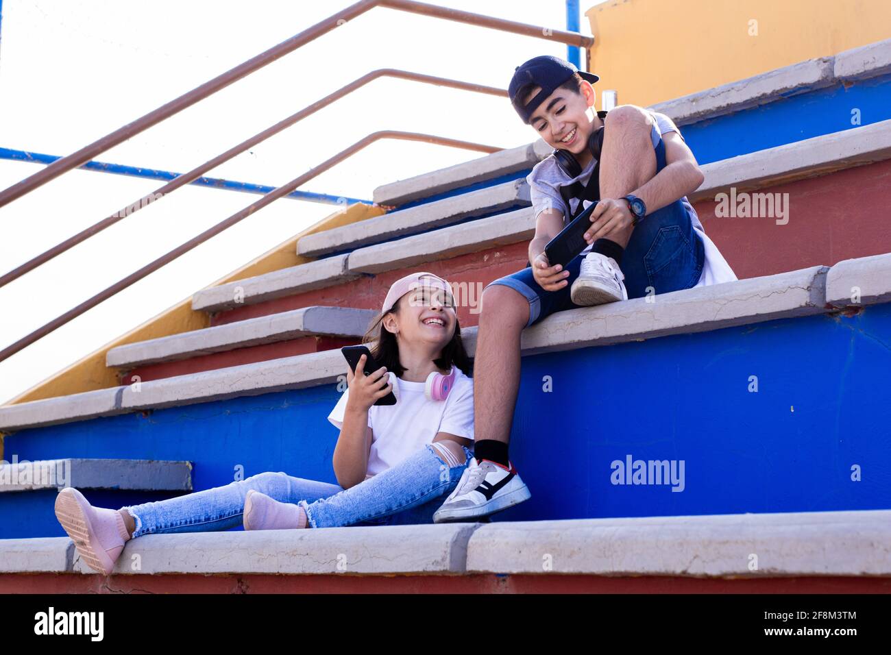 Due pre-adolescenti caucasici, molto sorridenti, con dispositivi mobili e cuffie, mostrando i loro telefoni cellulari l'uno all'altro seduti sui bleachers. Foto Stock