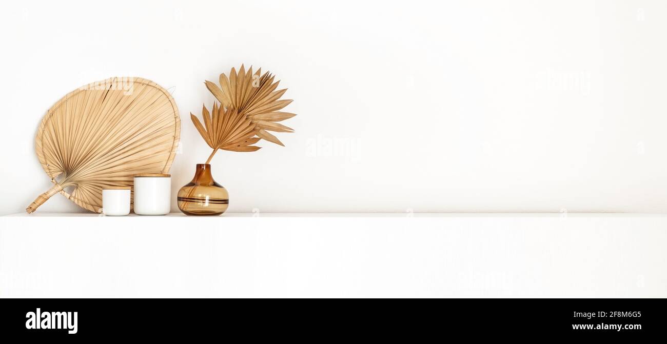 Testata con arredamento interno in stile boho costiero, un vaso di vetro marrone con ventilatori di palma essiccati e un cestino a parete intrecciato isolato su un ripiano bianco Foto Stock
