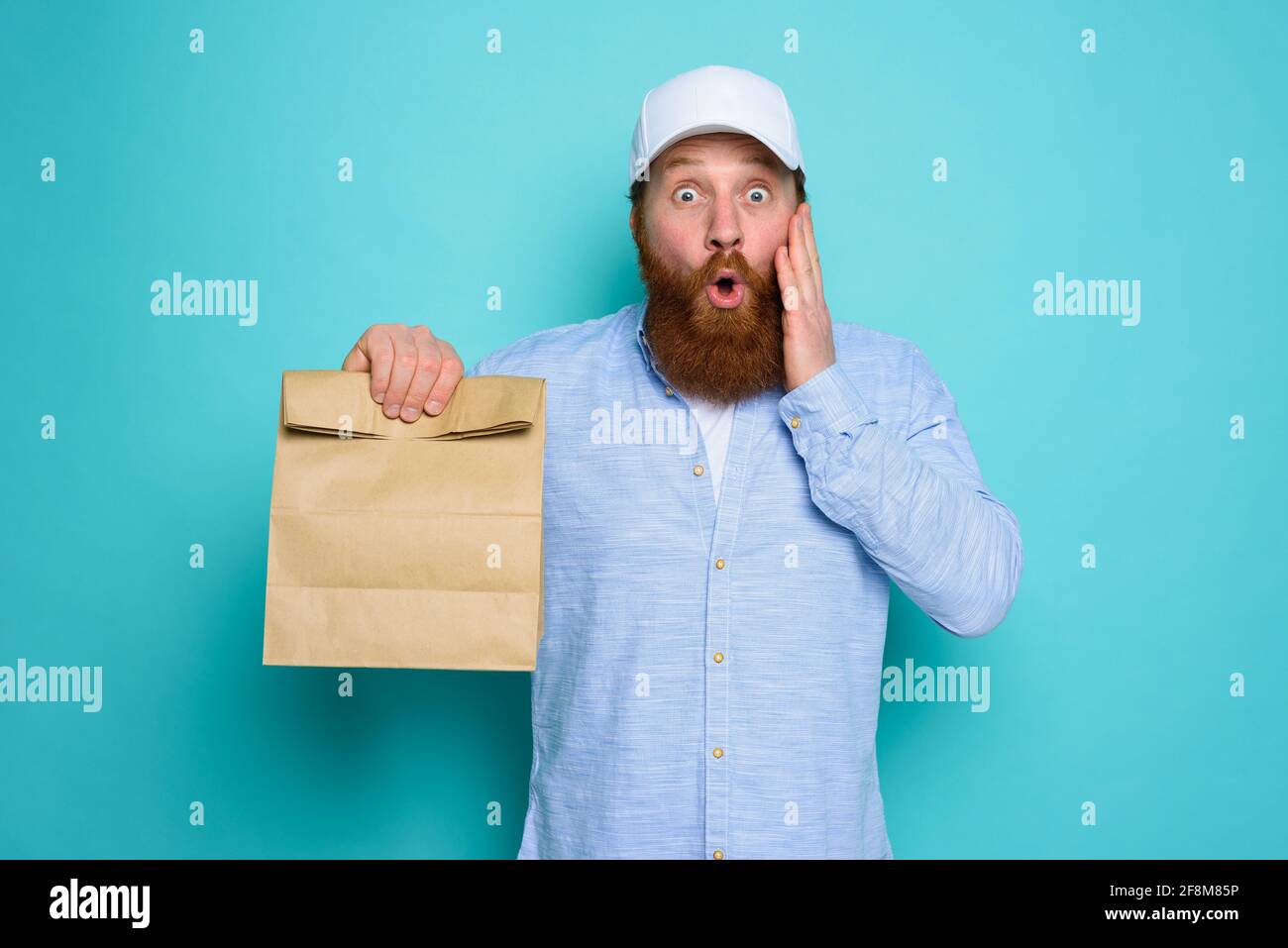 Deliveryman con espressione meravigliata pronto a consegnare il sacchetto con cibo. Sfondo ciano. Foto Stock