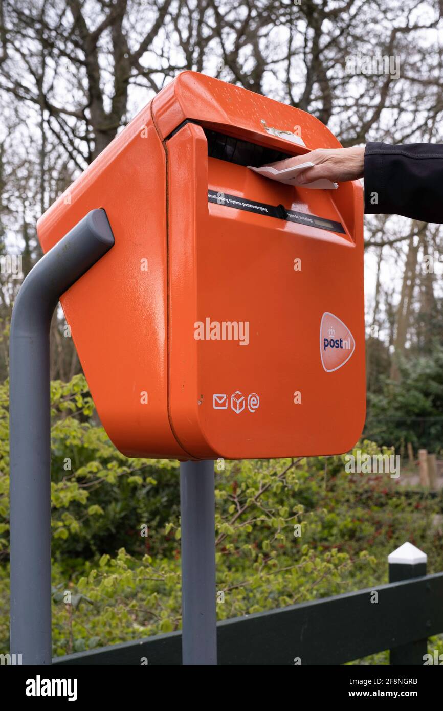La mano getta le lettere in una tipica scatola di lettere arancione  olandese, che si trova all'esterno in una strada lungo un parco Foto stock  - Alamy