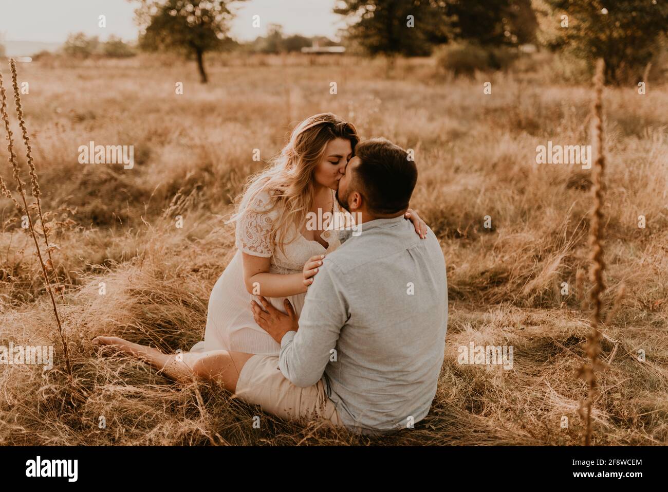 la moglie incinta si siede tra le braccia del marito e lo bacia e lo abbraccia. Felice famiglia riposo natura abbracci baci in estate al tramonto. Futura madre caucasica w Foto Stock