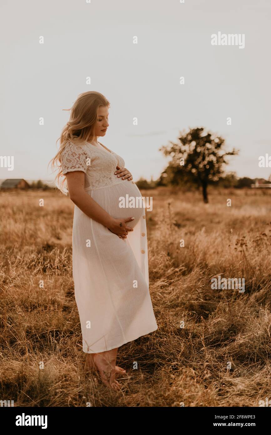 Caucasica giovane donna bionda incinta in cotone bianco abito in piedi prato camminare su erba secca in estate al tramonto natura. Madre-a-be la tiene Foto Stock